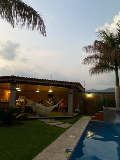The image shows an outdoor scene in San Francisco Lachigoló, Mexico, with a partially visible swimming pool on the right side. Beside the pool is a pathway made of light-colored rectangular stones and adjacent to the pathway is grass. An open-air structure is located to the left of the path, covered with a tiled roof and supported by brick columns. Inside the structure, there are several people seated at desks, illuminated by hanging lights, creating a potential workspace or gathering area. A white sail or awning-like structure is draped along one side of the open structure. The presence of palm trees on the left and top of the frame suggests a tropical or semi-tropical environment. The sky appears overcast. Mountains are visible in the distance.