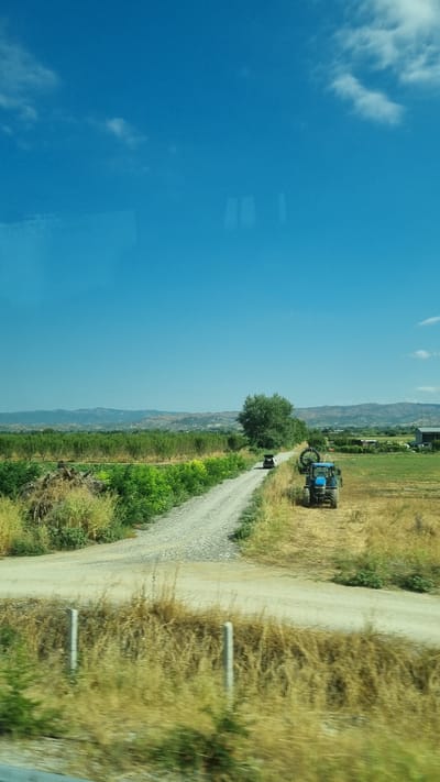 This image depicts a rural scene under a clear blue sky with scattered clouds. The foreground shows dry, yellowed grasses and two fence posts. A dirt road winds through the landscape, bordered by green vegetation and fields. Further down the road, a blue tractor is visible in a field of dry grass. In the middle ground, a dense green tree stands beside the road. Beyond the fields, a range of hills forms the distant horizon. The location is identified as Raxa, Greece. There are no people or overt actions visible in the scene. The overall impression is one of agricultural land and a peaceful, sunny day.