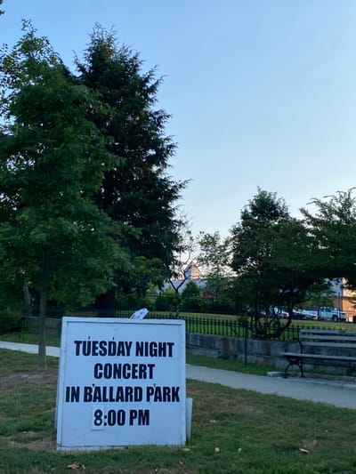 The image captures an outdoor scene in Ballard Park, Ridgefield, United States. A white sandwich board sign, positioned centrally in the foreground on green grass with scattered brown leaves, displays black text: "TUESDAY NIGHT CONCERT IN BALLARD PARK 8:00 PM". Behind the sign, a light-colored paved pathway extends diagonally to the right. To the right of the pathway, a park bench with dark metal supports and wooden slats is partially visible. A low stone wall topped with a black metal fence spans the midground horizontally. Various trees with green foliage are present, including a tall evergreen tree to the upper left of the sign and a deciduous tree immediately to its left. In the distant background, a white building with a dark roofline is partially visible through the tree canopy. The sky above is clear and light blue. No individuals or active events are present in the static scene.