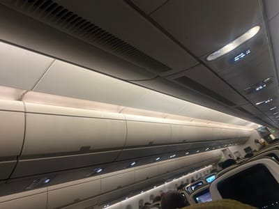 The image shows the interior of an airplane cabin. Overhead are closed overhead bins. The ceiling has air vents and lights with seatbelt signs illuminated. Rows of seats are visible with passenger entertainment screens. People are seated, and an airline attendant is visible in the distance. The location is not apparent from the image and there is no evidence to suggest the location is Addis Ababa, Ethiopia.