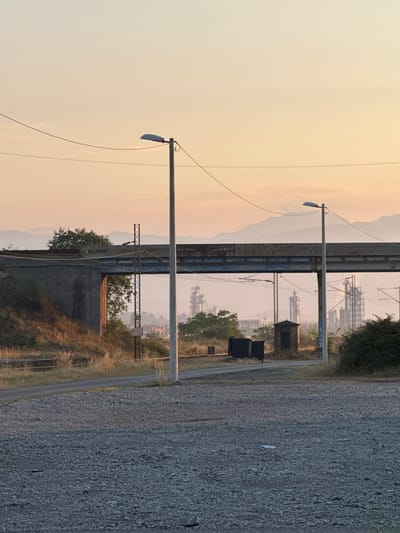 In Mojanovići, Montenegro, an industrial landscape is depicted during a soft dawn. A weathered concrete bridge, likely part of a railway infrastructure, spans across the frame, with its underside showing signs of rust and wear. Beneath the bridge, the skeletal structures of an industrial complex, possibly a refinery or power plant, rise into the hazy sky. Tall, slender chimneys emit plumes of steam, indicating ongoing industrial activity. A small, dark wooden structure, possibly a utility shed or signal box, sits near the tracks, flanked by two dark industrial bins. A single lamppost stands prominently in the foreground, its pole weathered and the lamp itself tilted. Power lines stretch across the scene, connecting to various poles. The ground in the foreground is covered in loose gravel, and a discarded plastic bottle lies on its surface. In the background, a series of soft, rolling hills or mountains fade into the pastel hues of the dawn sky. The overall atmosphere is one of industrial quietude under a gentle morning light.