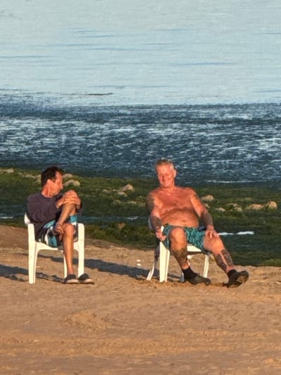 In the city of Shepelevo, Russia, two men are sitting on white plastic chairs on a sandy beach. The man on the left is wearing a dark t-shirt and patterned swim trunks. He is leaning forward with his arms crossed and his legs crossed. The man on the right is shirtless and wearing patterned swim trunks. He is leaning back with his arms resting on the arms of his chair. Both men appear relaxed. In the background, the sea stretches out to the horizon, with a band of green vegetation along the shore. The lighting suggests it is either early morning or late afternoon.
