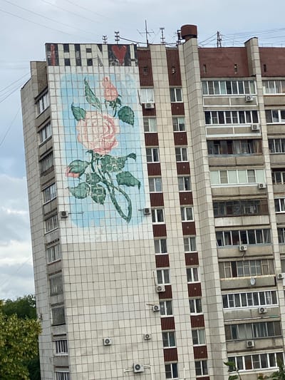 The image is a close-up, eye-level, exterior shot of a tall, multi-story apartment building. The building is constructed with light-colored square tiles. A large mural of a pink rose with green leaves is painted on a section of the building. The mural is set against a light blue, grid-like background. Above the mural, the top of the building features a sign with black and white stripes and a red heart. The sky is overcast with scattered clouds. The building has numerous windows, some with white frames and others with darker frames. Air conditioning units are visible on the exterior walls. To the left of the building, the tops of green trees are visible. The provided hints do not contain information about Yokohama, Japan.