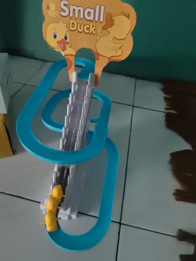 In Barengkrajan, Indonesia, a toy featuring a blue track with a vertical elevator mechanism is set up on a tiled floor. A yellow toy, resembling a caterpillar or segmented duck, is positioned on the track. Above the track, a decorative cardboard cutout displays the text "Small Duck" with cartoon ducks. The background is a solid teal-colored wall.
