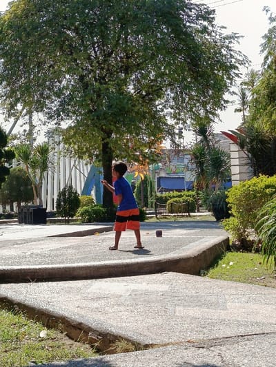 The image displays an outdoor scene in Asembagus, Indonesia, under clear daylight conditions. A single male child, dressed in a blue t-shirt and orange and black striped shorts, stands on a light-colored, multi-level paved pathway. He is positioned facing slightly right, with his left arm raised and bent at the elbow, appearing to hold or gesture with a small, light-colored object. His gaze is directed upwards and to the right. A reddish-brown, cuboid object rests on the paved surface to his right.
The background features a large, mature tree with dense green foliage. To the left of the tree, white vertical structures and a blue abstract sculpture are visible. To the right of the child, a dark metal park bench is situated amidst green shrubs and palm-like plants. Distant urban buildings, including one with a partially visible "HERBAL" sign, are discernible beyond the vegetation. Green grass areas are present in the foreground and to the right. The overall environment suggests a public park or urban green space.