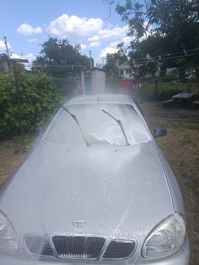 The image captures a silver Daewoo car being washed outdoors in a yard, likely on a sunny day given the bright sky and fluffy white clouds. The car's windshield and hood are covered in a thick layer of white foam, indicating the washing process is in progress. Water spray is visible on the windshield, and the windshield wipers are positioned upright. The car's headlights and grille are partially visible through the foam.

The setting is a residential backyard with a fence in the background. Several trees with lush green foliage are present, suggesting a warm season. Clothes are hanging on a clothesline strung between poles, with colorful clothespins holding them. A brick structure and part of a house are visible in the distance. The ground is covered in dry grass.

The overall impression is one of everyday life and domestic chores. There is no indication of specific emotions, but the scene suggests a casual, perhaps routine, car washing activity. The lighting and shadows indicate it's likely daytime, possibly midday or afternoon. There is no visible text in the image.
