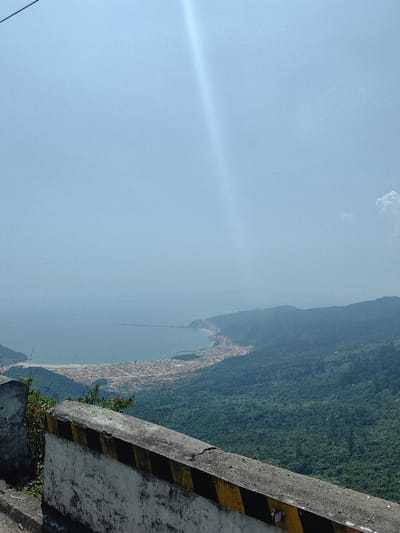 The image displays an expansive high-angle view of a coastal landscape within Hải Vân Ward, Vietnam, under a bright, hazy sky. In the immediate foreground, a weathered concrete road barrier or retaining wall is visible, featuring a white base with diagonal black and yellow stripes. Beyond the barrier, a dense green forested mountainside slopes down towards a large bay.
The bay's waters appear a pale blue-grey, extending towards the horizon where they meet a distant, hazy coastline. Along the curving shoreline of the bay, an extensive area of land appears developed or under development, characterized by numerous light-colored, rectangular plots or structures. A prominent, dark-colored pier or breakwater extends into the bay from the right side of the frame, partially enclosing an area of water. Several small, dark objects consistent with fishing boats are faintly visible in the water near the shore.
The background consists of multiple layers of lush, verdant green mountains and hills receding into a atmospheric haze. The sky is a light, clear blue, with a distinct, bright sun flare traversing diagonally from the upper left towards the center-right. A small, white cumulus cloud is present in the upper right quadrant. No people, specific actions, or interactions are discernible.