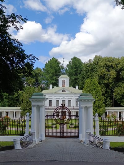 This is a full-shot, vertical, eye-level, outdoor photograph of a grand white estate with a large, ornate gate in the foreground. The estate building is a two-story structure with a green, domed cupola on top, and a balcony with a dark railing. The building has several windows and is surrounded by lush green trees. The gate is made of dark metal with intricate circular patterns and is flanked by large white pillars. A paved walkway leads up to the gate. A black metal fence with decorative elements runs along the sides of the property, with blooming red flowers visible in planters. The sky above is blue with scattered white clouds, suggesting a pleasant daytime with good weather. The scene evokes a sense of tranquility and historical grandeur. The location is identified as Ligachevo, Russia. There is no visible text in the image.