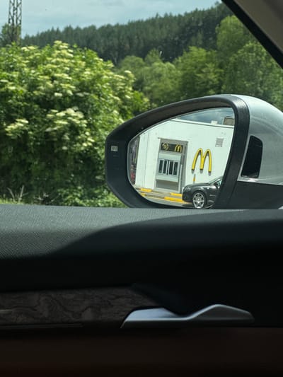The image, taken from inside a vehicle, shows a side-view mirror reflecting a McDonald's restaurant. The reflection captures the iconic golden arches logo and the building's white facade, along with a dark-colored car parked nearby. A portion of the restaurant's entrance with multiple windows is also visible. The mirror is framed by the vehicle's interior, with textured dark gray and brown panels visible. Outside the mirror, the reflection of trees, some with white blossoms, and a hazy sky can be seen. A metal structure, possibly part of a power line or fence, is visible in the lower left corner of the frame. The scene suggests a daytime setting, likely a sunny day due to the bright reflections. The location is identified as Studena, Bulgaria. There is no visible text in the image other than the McDonald's logo.
