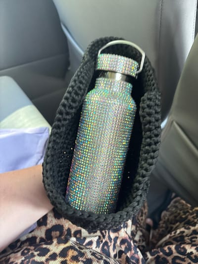 A person's arm is visible in the lower right corner of the image, resting on a pale purple cloth. The person is wearing a leopard-print garment. In the center of the image, a water bottle covered in iridescent rhinestones is nestled within a black crocheted bag. The bottle has a silver handle. The background suggests the interior of a car, with grey upholstery visible on the left and hints of what might be a car seat. The lighting appears to be natural, possibly daylight, given the reflections on the rhinestones. There is no visible text in the image.
