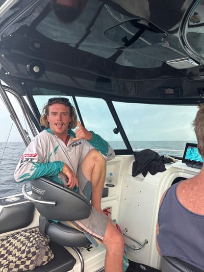 This image captures the interior of a boat operating offshore from North Wollongong, Australia. In the foreground, a young adult male with long blonde hair and sunglasses on his head is seated in a black upholstered swivel chair. He wears a white and teal long-sleeved fishing shirt with a "Makita" logo and grey board shorts. He is actively posing for the camera, extending his tongue and displaying a "shaka" hand gesture with his right hand. His right leg is bent with the foot elevated on the chair's armrest. To his right, the back of a second male's head and shoulders are visible; he has short brown hair and is wearing a dark tank top, facing forward towards the boat's bow.
The boat's cabin features white fiberglass panels and a black overhead console equipped with recessed speakers, lighting, and a digital display showing the number "12." A clear plastic water bottle is secured in a holder on the right side console. On the console in front of the second male, a tablet displays a blue and green topographical map or sonar reading. Folded black clothing rests near this tablet. The boat's windows reveal a view of the open ocean, characterized by dark grey-blue water under an overcast, light grey sky. A patterned, checkered blanket is draped over a seat or cooler on the left side of the frame.