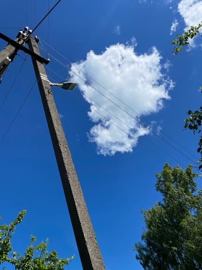 In Izborsk, Russia, a concrete utility pole angles upwards against a vibrant blue sky dotted with a single, fluffy white cloud. A streetlamp is attached to the pole, casting a subtle shadow. Power lines crisscross the frame, some taut and others slightly slack. To the left and right of the pole, lush green foliage from trees partially obscures the view. The image captures a clear day, with sunlight illuminating the scene.