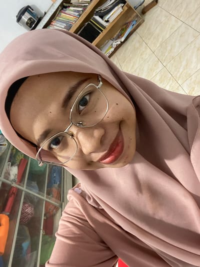 This is a selfie taken by a woman wearing a light pink hijab and glasses. She is looking directly at the camera with a slight smile. Her lips are painted with a shimmery red lipstick. Her eyes are a striking blue. The background is blurry, but appears to be indoors, possibly a room with shelves filled with books and other items. There is a wooden shelf with books on the left side of the image, and a glass-fronted cabinet filled with colorful items on the right. The lighting suggests it is daytime. The overall mood of the image is casual and friendly. The location context is Tanjung Gading, Indonesia.