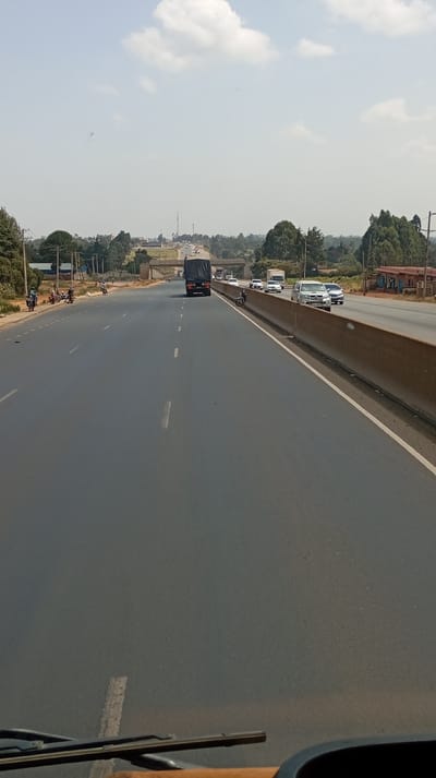 The image displays a multi-lane highway in Sigona ward, Kenya, captured from a vehicle's dashboard perspective on a clear to partly cloudy day. The dark asphalt road extends into the distance, marked by dashed white lane lines in the direction of travel. A concrete barrier separates traffic flowing in the opposing direction on the right, where a white van, a dark SUV, and other cars are visible, some in motion and one motorcycle parked on the shoulder.
On the left side of the camera's travel direction, a wide shoulder or service road is present, where multiple motorcycles are parked. Several individuals are standing near these motorcycles. Further along this side, utility poles are visible, and green trees line the road, interspersed with small buildings in the middle distance. A large black semi-truck is observed in the left lane of the highway, traveling away from the camera. Beyond the truck, an overpass spans the highway, and a tall communication tower is faintly visible in the far distance to the left. On the right, past the opposing lanes, green trees and a single-story building with a red roof are visible closer to the camera. The sky above is pale blue with scattered white clouds.