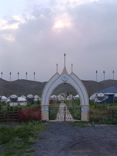 The image depicts an entrance gate to an ethno-complex in Naryn, Kyrgyzstan. The gate is an ornate arch with cream and white stripes, topped with pointed finials. It is flanked by tall poles with pointed tips, reminiscent of spears. Beyond the gate, a pathway leads into a village of yurts, traditional nomadic dwellings, situated on a gentle hillside. In the background, rolling hills are visible under a cloudy sky, with the sun peeking through the clouds, suggesting it is late afternoon or early evening. The weather appears overcast, but not raining.
Two signs are visible on the gate. The left sign has Cyrillic text that reads "Этно Комплекс Им. Алмаз Акматова" (Ethno Complex named after Almaz Akmatov). The right sign is partially obscured but also appears to be in Cyrillic and likely contains similar information.
The overall scene evokes a sense of history, tradition, and the nomadic lifestyle of Central Asia. The setting is rural and serene, with the yurts and natural landscape contributing to a peaceful atmosphere. There are no people visible in the image, highlighting the structures and the environment. The use of metal circular decorations, possibly shields, on poles further emphasizes the cultural theme. The ground in front of the gate is covered in gravel, and patches of green grass and vegetation surround the area.
