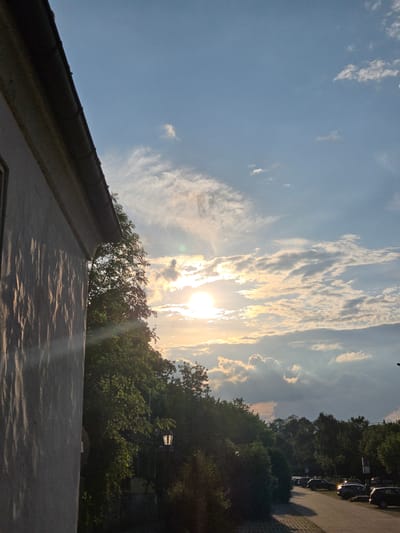 The image depicts a sunset scene in Ansbach, Germany. In the foreground, a portion of a building's white textured wall is visible on the left, with diagonal shadows cast upon it. Adjacent to the building, a dense canopy of green trees dominates the middle ground. A streetlamp with a lit bulb is partially visible among the trees. Further back, a parking lot is lined with several dark-colored cars. The sky is a blend of blue and white, with scattered clouds illuminated by the bright, low-hanging sun. The overall atmosphere is one of a tranquil evening.
