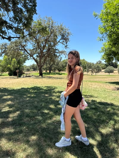 A young girl with long, light brown hair is standing on a grassy field, looking at the camera with a slight smile. She is wearing a brown short-sleeved shirt, black shorts, and white sneakers with grey accents. She is holding a light blue jacket and a pink bag with a cherry pattern over her left arm. Behind her, there are several trees with green leaves, and a picnic table with a trash can nearby. The sky is clear and blue. The location appears to be a park or a picnic area in Elverta, California, United States.