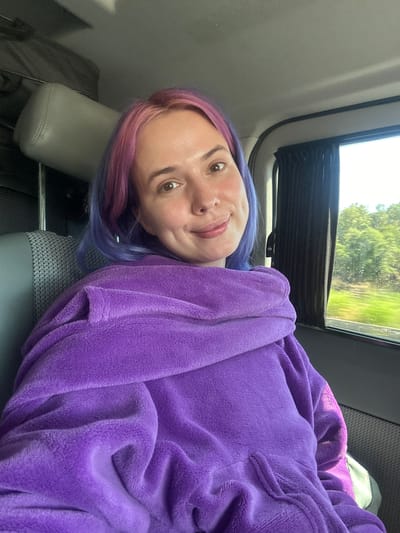 The image shows a woman with dyed pink and blue hair lying in what appears to be a vehicle. She is wearing a purple plush garment. A window is visible in the background, showing trees and greenery. The location is potentially Le Muy, France, as per the prompt's instructions, though no specific elements in the image directly confirm this.