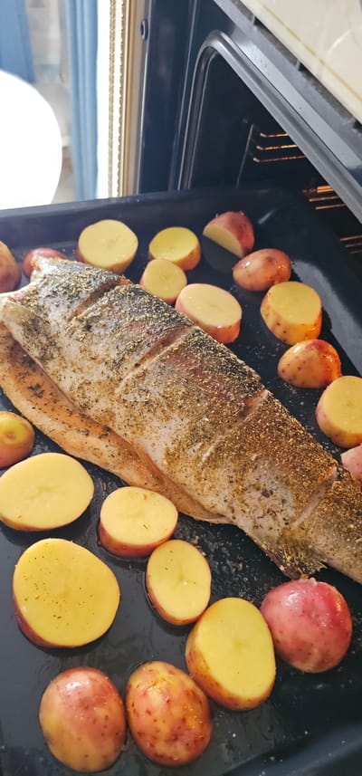 A whole roasted fish with seasoned skin lies on a black baking sheet. The fish is a prominent feature, positioned centrally and taking up a significant portion of the frame. Surrounding the fish are numerous halved red-skinned potatoes, their yellow flesh exposed. Some potatoes are whole, while others are cut in half, revealing their starchy interiors. The baking sheet appears to be placed inside an oven, as suggested by the presence of heating elements visible at the top of the image. The lighting appears to be coming from inside the oven, creating a warm and slightly shadowed atmosphere. The overall scene depicts the preparation or cooking of a meal, with the fish and potatoes ready to be roasted. No people are visible in the image. The setting is domestic, likely a kitchen, and the activity is clearly cooking. There is no visible text.