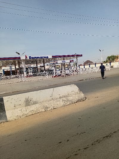 This image depicts a street scene in Sharada, Nigeria. A Danmarna fuel station occupies the left mid-ground, featuring a multi-pump setup beneath a red, white, and blue striped canopy. A sign reading "VESEL" is visible above one pump island. Multiple vehicles are present at the pumps, indicative of ongoing service. A white metal barrier delineates the station property. In the immediate foreground, a weathered concrete median barrier divides the asphalt roadway. To the right, a single individual, male, dressed in dark clothing, walks away from the camera along the road's shoulder. The environment is illuminated by direct sunlight, casting shadows. The sky is clear blue with faint overhead power lines. Distant buildings and trees line the horizon on the right.