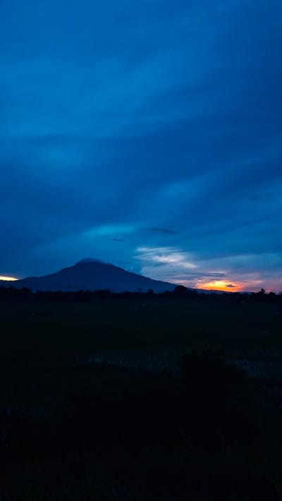 A solitary, dark mountain peak rises from a silhouette of trees in the middle ground of the image, shrouded in a light cloud near its summit. The sky above is a deep, rich blue, with streaks of lighter blue and grey clouds. To the right of the mountain, a sliver of orange and yellow light pierces through the horizon, suggesting a sunset or sunrise. The foreground is a dark, indistinct field, possibly a rice paddy, with some vegetation visible. The overall scene is serene and atmospheric, indicative of dusk or dawn in a rural setting. The image appears to depict the natural landscape near Baksukon, Indonesia, where such mountainous terrain and agricultural fields are common.