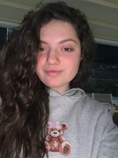 The scene is located in Piperkov Chiflik, Bulgaria. A female subject is centrally positioned, facing directly forward. She has medium-dark brown, curly hair that cascades over her left shoulder and partially covers the front of her torso. Her skin tone is fair, and she has brown eyes, a neutral closed-mouth expression, and looks directly at the camera. She is wearing a light grey hooded sweatshirt. On the left side of her chest (viewer's right), the sweatshirt features a graphic of a brown teddy bear with a pink bow. The background is out of focus, indicating a shallow depth of field. It depicts an outdoor or semi-enclosed nighttime environment. To the subject's left (viewer's right), faint distant lights are visible against a dark sky, along with partially visible, light-colored outdoor patio furniture, specifically a slatted chair or lounger. A light-colored ceiling or overhang structure is visible at the top of the frame.