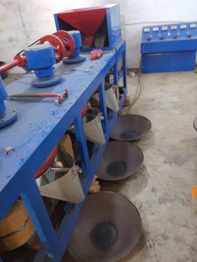 This forensic image details an industrial processing facility in Olomoda, Nigeria. The central subject is a long, blue-framed machine. Its upper structure incorporates a red hopper, multiple red pulleys, and associated blue cylindrical bearing housings. A red-handled tool rests on the machine's frame. Below the machine's operational surface, six light-colored fabric chutes descend into corresponding dark, shallow, circular collection pans on the concrete floor. Each pan contains a conical accumulation of dark granular material. In the background, a blue control panel unit is mounted on a raised platform, featuring five visible gauges and knobs; one indicator illuminates with a faint yellow light. The environment consists of light-colored walls and a stained concrete floor. No personnel are present.