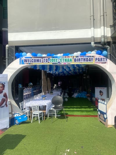 The scene is a birthday party entrance decorated with blue and white balloons in Bonadikombo, Cameroon, located at coordinates 4.048 latitude and 9.218 longitude. The party is for a child named Joel-Ethan. The entrance is marked by a banner that reads "Welcome to Joel-Ethan Birthday Party." The entrance is an archway made of white plaster, covered with blue and white balloons. The entrance leads into a courtyard decorated with white tables, chairs, and green artificial grass. There is a large poster on the right side of the image that reads "Joel-Ethan @ 3" depicting a cartoon boy. There is a lone black chair in the foreground and a cartoon car on the left side of the image. A young child is pictured on a poster on the left side of the image. Several people are visible in the background, but it is difficult to discern what they are doing. The scene is sunny and bright, with the image taken during the day.