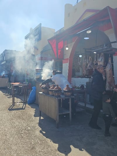 The scene depicts an active outdoor market or street food area in the city of Boufakrane, Morocco, under bright daylight with strong shadows. In the midground, a man in a white shirt and dark pants, wearing a dark cap, is actively tending to a cooking station. This station consists of a metal table holding at least five traditional clay tagine pots with conical lids, from which significant plumes of white smoke are rising. A blue plastic bag rests on the ground next to this station. To the left, several square metal tables and chairs are arranged, suggesting an outdoor eating area.

On the right side of the midground, a man in a dark jacket and a red baseball cap stands, observing the scene, with a young child in dark clothing standing beside him.

In the background, a building with a red and white striped awning shelters a butcher's stall on the right, where numerous animal carcasses are visibly hanging from hooks. To the left, another building features a prominent blue sign with white Arabic script. Further left, additional cooking activity with smoke is visible. The ground is a light-colored, coarse paved surface.