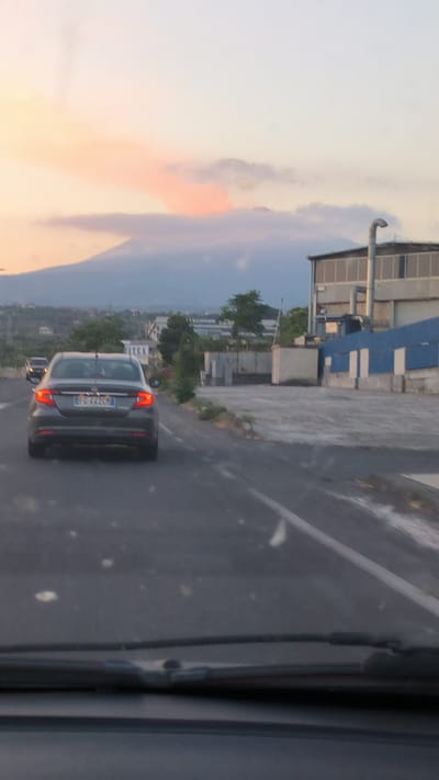 The image captures a street scene in Belpasso, Italy, with a prominent volcano in the background. A dark gray Fiat Tipo sedan is the primary subject, driving on an asphalt road. The car's license plate is visible, reading "FG 442CN". In the distance, several industrial buildings are present, including one with a corrugated metal exterior and another with blue panels. The sky is partly cloudy, with a hazy, orange glow suggesting either sunset or a volcanic eruption plume. The foreground shows the interior of a vehicle, with a windshield and wipers. The overall atmosphere is somewhat hazy and subdued, possibly due to the atmospheric conditions or time of day.