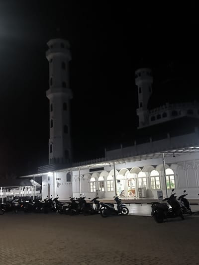 In Barat Yaman, Indonesia, at night, a mosque with white minarets and a white facade stands under a dark sky. The mosque features arched windows and decorative relief work on its walls. In front of the mosque, a line of parked motorcycles fills the paved area. The scene is illuminated by artificial lighting from the mosque's interior and exterior. No people are prominently visible in the image.