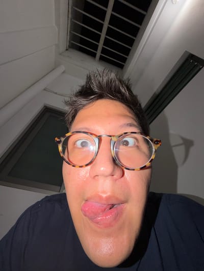 A person with short, dark hair and tortoiseshell glasses makes a silly face, sticking out their tongue. They wear a dark blue or black shirt. The shot is taken from below, at an extreme angle. Visible in the background are a window with dark vertical bars, a dark rectangular window, and a linear ceiling light fixture. A faint, unclear shadow is cast above the person.