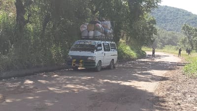 The image shows a white van parked on a dirt road in Rutshuru, Democratic Republic of Congo. The van is overloaded with bags and other cargo on its roof. There are two people walking on the road in the distance. The van is parked on the right side of the road, with a line of trees on the left side of the road. The road is dusty and there are shadows from the trees. The van is facing forward and the road appears to curve slightly to the left. The terrain is hilly, and the road is surrounded by trees and vegetation. The image is taken during the day and the sun is shining. The van has the words "For all Land" painted on the side in green and white, and a license plate reads "K 611 AC". The road is relatively empty with only a few people visible in the distance. The surrounding environment suggests a rural setting in Africa.