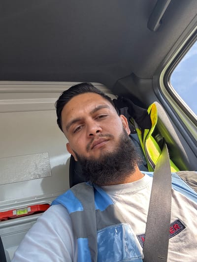 A man is taking a selfie inside a vehicle. He is looking directly at the camera with a neutral expression. He has dark hair, a beard, and brown eyes. He is wearing a white t-shirt and a blue and gray safety vest. A seatbelt is visible across his chest. In the background, there are some items stored in the vehicle, including a red level. The interior of the vehicle appears to be a cargo area, possibly a van. The lighting suggests it is daytime. There is no specific information in the image to indicate the location is the city of Namur, Belgium.
