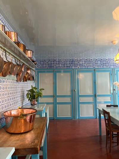 The image shows a kitchen inside a house in Giverny, France. The kitchen is located at the coordinates 49.075 latitude and 1.534 longitude. The kitchen is decorated in a French country style, with blue and white tile walls, a white ceiling, and a red tile floor. The kitchen has a wooden countertop and cupboards, with several copper pots hanging on a rack on the wall above them. A large copper pot is resting on the counter. The walls are covered in blue and white tiled patterns. There are three blue-painted doors with white trim. A white table with a chair is visible in the background.  The light source in the room is an open doorway from another room, which appears to be connected to the kitchen. The kitchen appears to be empty of people.