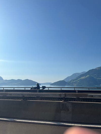 The image depicts a daytime scene over Lake Lucerne (Vierwaldstättersee) in Beckenried, Switzerland, viewed from a motorway bridge. The sky, occupying the upper two-thirds of the frame, is clear and light blue. The midground features the calm, blue waters of Lake Lucerne, bordered by multiple layers of forested mountains receding into the distance.
On the bridge, in the left midground, a single individual is visible, operating a dark-colored motorcycle. The rider is wearing a dark helmet and appears in silhouette, facing towards the left. The motorcycle is equipped with a dark top case or luggage on its rear.
The foreground displays the structural elements of the bridge. A substantial concrete barrier or wall runs horizontally across the lower portion of the image, topped by a metal guardrail system. The dark asphalt road surface is visible at the very bottom of the frame. The scene is illuminated by bright sunlight, casting the foreground elements and the motorcycle into partial silhouette against the bright lake and sky.