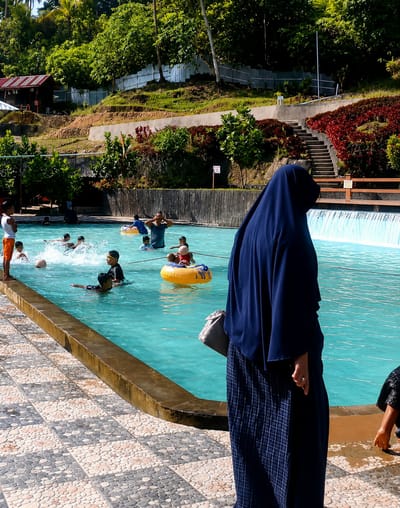 The image depicts a lively scene at a water park in Silaiyajae, Indonesia. The focal point is a large, turquoise swimming pool filled with a diverse group of people, primarily children, enjoying various water activities. In the foreground, a woman dressed in a dark blue hijab and a plaid skirt stands facing away from the camera, holding a light gray purse. Her hand, adorned with a ring, is visible at her side. The pool itself is surrounded by a patterned tiled walkway and a brown-edged pool border.
Several children are actively engaged in the water. Some are swimming, while others are utilizing inflatable rings. One child is seen floating in a yellow inner tube. In the background, a man in a dark blue shirt is partially submerged in the water, with his arms raised. Further back, other individuals can be seen swimming and playing in the pool.
The environment surrounding the pool suggests a lush, tropical setting. Dense green trees and vegetation cover the hillside behind the water park. A small, red-roofed building is nestled on the hillside, and a set of concrete stairs leads up the embankment. A metal fence runs along the top of the hill. The water park appears to be built into the natural landscape, with terraced areas and strategically placed foliage. The overall atmosphere is one of recreational enjoyment on a sunny day.