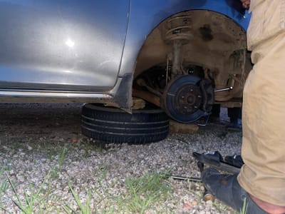 The image shows a person's legs and torso from the waist down, wearing khaki pants and black work boots, as they are working on a car. The car's wheel is removed, revealing the brake assembly and suspension system. A spare tire is lying on the ground next to the car. The scene is set outdoors, with gravel and dry grass visible on the ground. The lighting suggests it might be daytime, possibly late afternoon or early evening, given the slightly dim and somewhat diffused light. The environment appears somewhat unkempt, with scattered debris and vegetation. The overall impression is one of vehicle maintenance or repair in a rural or roadside setting. There is no visible text in the image. The activity is clearly focused on changing or inspecting the car's wheel. The scene is not specific enough to infer emotions, weather conditions beyond what can be deduced from the lighting, or a precise time of day. The location context "Gbessa, Nigeria" provides a geographical setting, but no specific visual cues within the image directly confirm this location.