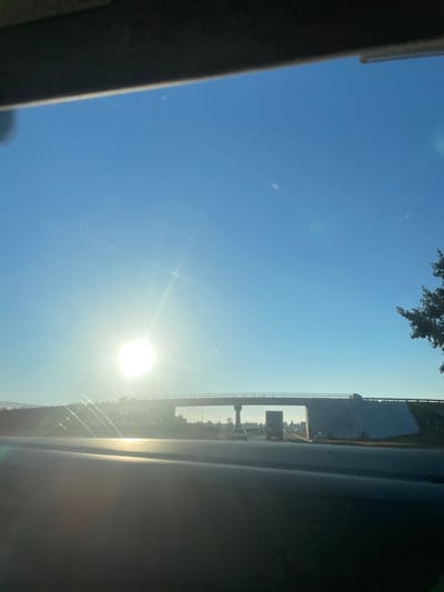 The scene depicts a bright, clear day on a highway within Amozoc de Mota, Mexico, captured from the interior of a vehicle looking forward through the windshield. The upper section of the image is dominated by a deep blue sky. The sun, positioned prominently to the left of the frame's center, emits intense light and significant lens flares. A concrete overpass, supported by pillars and featuring a visible railing, spans horizontally across the mid-ground. Beneath this overpass, a multi-lane roadway is visible, hosting vehicular traffic including at least one large truck and multiple passenger vehicles. The immediate foreground shows the dark, curved surface of the vehicle's dashboard or interior trim, reflecting some of the sunlight. Sparse vegetation and distant structures are visible along the road margins, indicating a semi-urban highway environment.