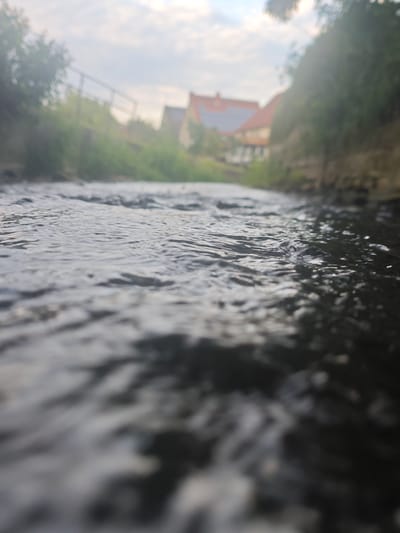 The image shows a low-angle view of a small, flowing body of water, likely the Emmer river in Lügde, Germany. The water's surface is disturbed by ripples and small waves. The banks of the watercourse are visible, with greenery and some trees or bushes. In the background, out of focus, are buildings with red roofs, suggesting a residential area. The sky is partly cloudy and bright, indicating daytime. The perspective is from the water level, looking upstream towards the buildings and landscape.