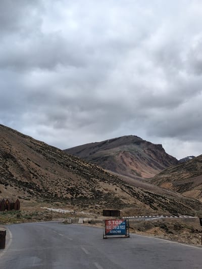 The image depicts a desolate, mountainous landscape under a heavily overcast sky. A paved road curves through the scene, with steep, barren hillsides covered in sparse vegetation on either side. In the foreground, a temporary road barrier with a "STOP POLICE CHECK POST SERCHUU" sign is positioned on the right side of the road. To the left of the road, further in the distance, are a few small, rust-colored structures that appear to be abandoned or storage units. The overall impression is one of isolation and rugged terrain, with the police checkpoint suggesting a remote, controlled access point. The specific location of Dhar Chharp Samba, India, is not visually indicated within the image itself but is provided as contextual information.