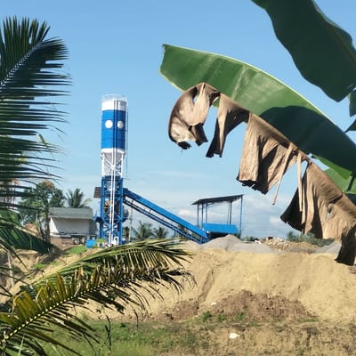This media file captures an industrial scene, likely a concrete batching plant, under a bright, clear blue sky in Seuneubok, Indonesia. The central subject is a tall, blue and white cylindrical tower, characteristic of a cement silo, supported by a metal framework. A blue conveyor belt system extends diagonally from the lower part of this tower towards the right, leading to mounds of aggregate materials such as sand and gravel piled on the ground.
The foreground and sides of the image are framed by lush tropical vegetation. On the left and bottom, vibrant green palm fronds are visible, while large banana leaves, some green and others dried and brown, hang from the top and right, partially obscuring the view. In the background, other palm trees and a simple building with a light-colored roof can be seen.
The scene suggests an ongoing industrial operation for producing construction materials. The bright sunlight and absence of clouds indicate a clear daytime setting, likely mid-morning or afternoon, with strong natural lighting. A circular emblem or logo is visible on the blue section of the main cylindrical tower, though the text within it is not decipherable. No people are visible, and the scene is devoid of overt activity, presenting a static view of the industrial site within its natural surroundings.