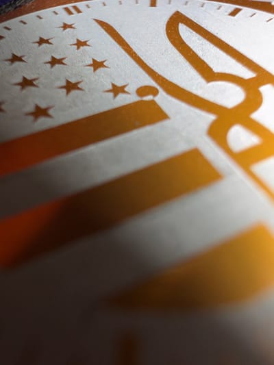The image is a close-up, angled shot of what appears to be a part of a medal or commemorative coin. The surface is predominantly silver-toned with raised, glossy gold elements. These gold elements include what look like stripes resembling the American flag and several small, five-pointed stars. There are also larger, stylized gold markings in the upper right quadrant, possibly representing numerals or a crest. The lighting creates significant shadows and highlights, obscuring some details and adding a dramatic effect to the texture of the surface. There is no indication of a specific location in Grapevine, United States of America.
