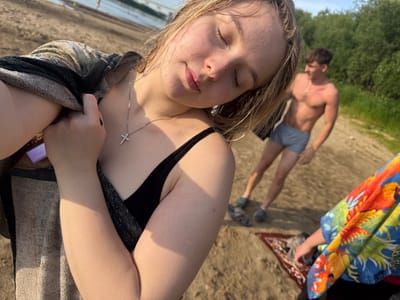 The image shows two young people on a sandy beach. The foreground is dominated by the face and upper body of a young woman who appears to be lying down, with her eyes closed. She is wearing a black bikini top and a silver necklace with a cross pendant. A towel is draped over her legs. In the background, a young man is visible, standing and looking away from the camera. He is wearing light blue swim trunks and appears to be shirtless. A colorful beach towel with a fish motif and a small rug are also present on the sand. The setting suggests a sunny day, likely in the late afternoon or early evening, given the warm, golden light and shadows cast on the sand. The presence of trees and a body of water in the distance suggests a natural setting, possibly near a lake or river in Ryabinino, Russia. The overall mood is relaxed and casual, indicative of a day spent outdoors enjoying the summer weather. There is no visible text in the image.