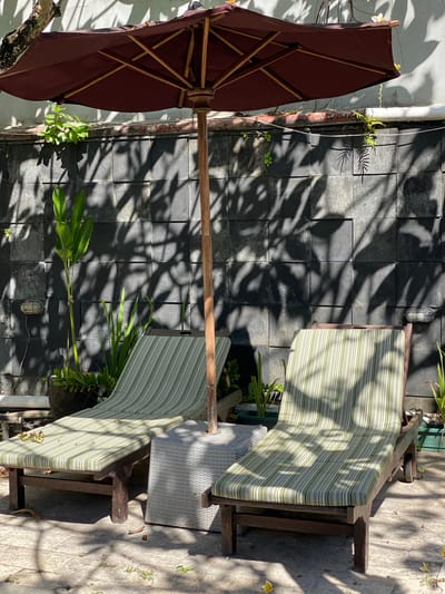 A close-up, eye-level, outdoor shot shows two empty lounge chairs with green and white striped cushions in front of a dark stone wall. The wall is textured and features shadows cast by overhead foliage. A large, dark red umbrella with a wooden pole is positioned between the chairs, providing shade. In the foreground, the ground is paved with stone tiles, and scattered fallen leaves are visible. Several potted plants with green foliage are situated behind and between the lounge chairs. To the left, a small, weathered light fixture is mounted on the wall. The scene suggests a tranquil outdoor seating area, possibly a garden or patio in a warm climate. The lighting indicates it is daytime, with strong sunlight casting distinct shadows. This setting is consistent with a resort or private residence in Denpasar Barat, Indonesia, where such outdoor amenities are common.