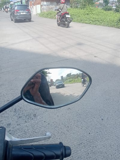 In a street scene in Sawotratap, Indonesia, a person riding a motorcycle is captured. The image is taken from the perspective of another motorcycle rider, looking into their side mirror. The mirror reflects the rider's hand and arm, which are wearing a dark garment. Also visible in the mirror is the road ahead, with a dark-colored car in the distance. Beyond the car, another motorcycle with a rider wearing a red helmet and a light-colored jacket is visible, traveling in the same direction. The road surface is asphalt, and the surrounding environment includes sparse vegetation and buildings on the side of the road. The weather appears to be clear and sunny, casting shadows on the road.