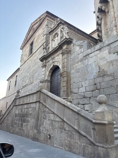 The image displays the main entrance of the Convento y Museo de Santa Teresa, located on Plaza de la Santa in ÁVila, Spain. The primary subject is a large, multi-story building constructed from rough-hewn grey stone blocks. A symmetrical, double-winged stone staircase ascends from ground level to the arched main entrance. The staircase features stone balustrades with spherical finials topping the newel posts. The entrance itself is a dark, arched wooden door adorned with a grid pattern of metallic studs.
Above the doorway, an ornate stone portal includes a central niche housing a white statue of a robed figure, presumably a saint. This central statue is flanked by two additional white statues, each standing on a pedestal positioned on the entablature directly above the archway. A triangular pediment crowns the central portion of the facade. To the right of the main entrance, the stone wall continues, and an architectural element with a corbelled design is visible in the upper right quadrant. The sky is clear and light blue. In the lower-left corner of the frame, a partial view of a vehicle's side mirror is visible, reflecting a portion of a human head. No other individuals are present. The scene is illuminated by natural daylight.