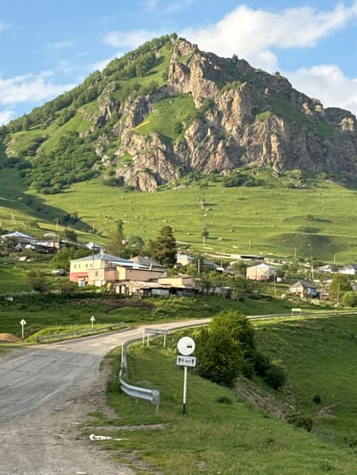 A paraglider with a yellow wing is visible against the backdrop of a steep, rocky mountainside covered in lush green grass. The mountain slopes gently down to a village with several houses nestled amongst the greenery. A paved road winds through the village, with some houses situated close to the road. Several white directional signs are posted along the road. One sign features a circle with an arrow pointing right and the text "6 км" which translates to "6 km". Another sign displays Russian text, which appears to be "НИЖНЯЯ МАРА" (Nizhnyaya Mara), indicating the village's name. The sky is blue with scattered white clouds. The overall scene suggests a bright, sunny day in a mountainous region.