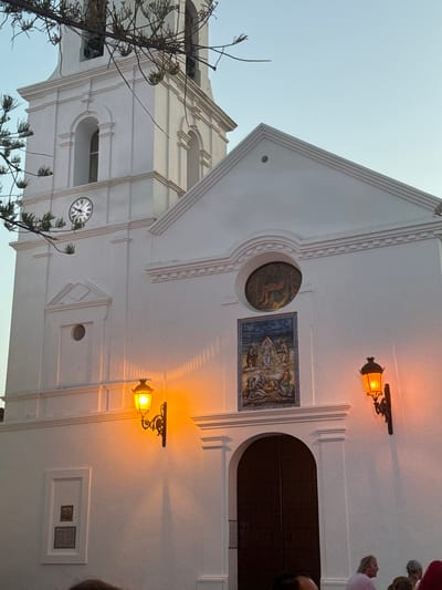 The image shows the exterior of the Church of El Salvador in Nerja, Spain, at dusk. The white facade of the church is illuminated by two ornate, wrought-iron lanterns with orange light, casting shadows on the wall. A large, arched wooden door is centered on the lower part of the facade. Above the door, a rectangular tiled panel depicts a religious scene. Higher up, a circular window contains a mosaic of what appears to be Christ's entombment. To the left, a church tower with a clock face is visible, with branches of a tree obscuring part of it. The sky above is a clear, pale blue. In the lower portion of the image, the heads and shoulders of several people are visible, suggesting a gathering or event.