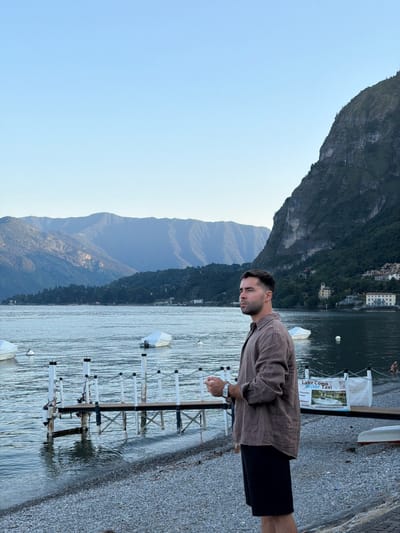 A man stands on a pebble beach in Loveno, Italy, looking out at a calm lake. The man is wearing a brown linen shirt and black shorts. In the background, there are mountains and a few boats on the water. A wooden pier with white railings extends into the lake, and a sign for "Lake Como Water Taxi" is visible. The sky is clear and blue.